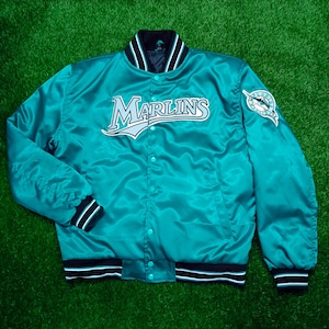 May include: Teal satin jacket with the word "Marlins" in white and black lettering across the chest. The jacket has a button-up front, black and white striped trim, and a team logo patch on the sleeve.