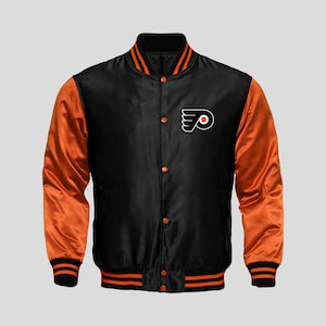May include: A black satin varsity jacket featuring vibrant orange sleeves and a black and orange Philadelphia Flyers logo on the left chest. The jacket includes a black ribbed collar, cuffs, and hem with orange stripes, plus a front snap button closure.