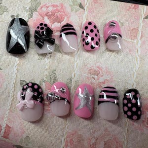 May include: A collection of ten artificial nails with various designs. The nails feature black, pink, and white bases with silver star, polka dot, bow, and striped patterns. Some nails have rhinestones.