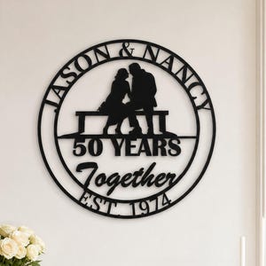 May include: A circular black metal wall sign featuring the names JASON & NANCY at the top, a silhouette of a couple sitting on a bench in the center, and the text 50 YEARS Together EST. 1974 below the bench, all enclosed within a thin circular frame.