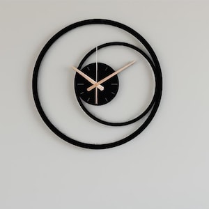 May include: A contemporary black wall clock with a distinctive design. The clock has a black circular frame and a smaller black circular clock face. The clock hands are light brown. It is mounted on a white wall.