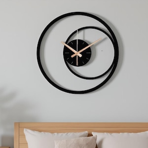 May include: A modern black wall clock with a unique spiral design. The clock features a black face with gold-colored hands and a second hand. The clock is mounted on a light gray wall above a bed with pillows.
