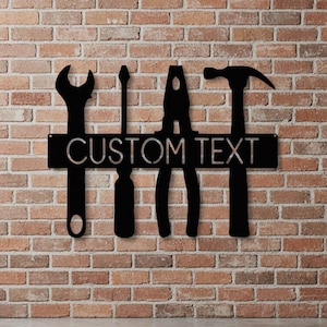 Puede incluir: Un letrero de pared de metal negro con siluetas de una llave inglesa, un destornillador, unos alicates y un martillo dispuestos horizontalmente. Una barra rectangular central cruza las herramientas y muestra el texto CUSTOM TEXT en negrita.