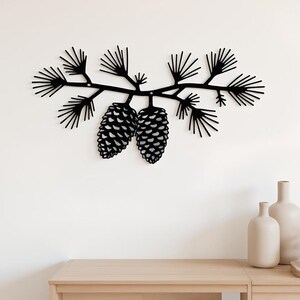 Op de afbeelding: Zwarte metalen wandkunst met een dennentak met twee gedetailleerde dennenappels. Het kunstwerk is bevestigd aan een witte muur boven een houten kast, waardoor een natuurlijke, rustieke esthetiek ontstaat. Het ontwerp is een minimalistische weergave van de natuur.