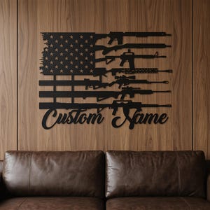 May include: Black metal wall art depicting the American flag with rifles replacing the stripes. The flag design includes stars and the text "Custom Name" in a cursive font. The artwork is mounted on a wooden wall.