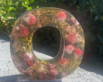 Könnte beinhalten: Ein klarer Harzbuchstabe O mit eingebetteten getrockneten Blumen und Grün. Der Buchstabe ist hellgelb mit rosa Blüten und grünem Laub.