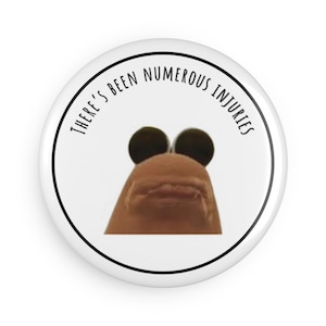 May include: A white badge with a black border and the text "THERE'S BEEN NUMEROUS INJURIES" in black. The badge features a close-up image of a finger puppet with large black eyes and a small mouth.