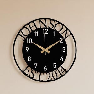 Puede incluir: Un reloj de pared negro y circular con el nombre "JOHNSON" recortado en el borde. La esfera del reloj es negra con manecillas doradas y números blancos. El texto "EST 2014" está en la parte inferior.