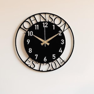 May include: A black wall clock with the name "JOHNSON" cut out around the circular frame. The clock face is black with gold-colored hands and numbers. The text "EST 2014" is below the clock face.