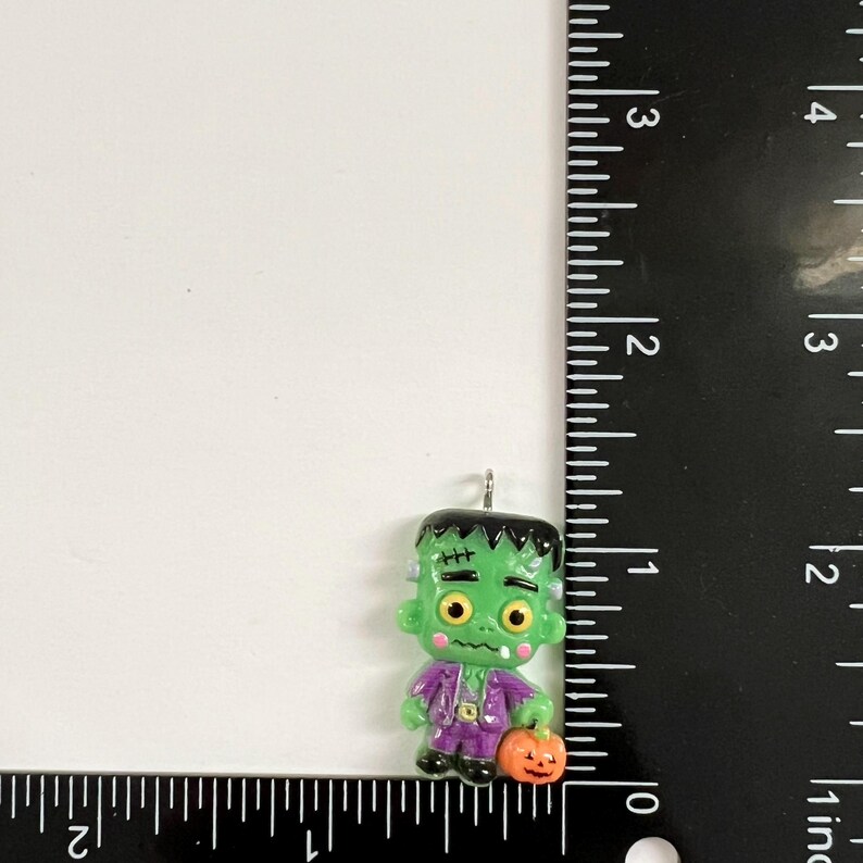 May include: A green Frankenstein monster charm with black hair, yellow eyes, and a purple suit, holding a small orange pumpkin. A black ruler is positioned along the right side and bottom, showing the charm is approximately 1.5 inches tall.