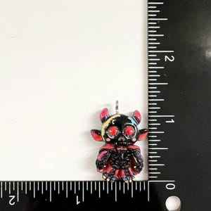 May include: A small black devil pendant with bright red eyes and colorful accents, featuring a skeleton-like body and a cape. The pendant is positioned next to a black ruler, showing it measures approximately 1.25 inches in total height.