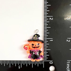 May include: A pumpkin-shaped charm wearing a black witch hat and cape, holding a red and green lollipop. The charm features a silver metal loop at the top and is positioned next to a black ruler, showing a height of approximately 1.5 inches.