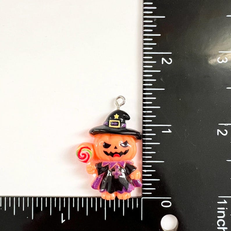 May include: A pumpkin-shaped charm wearing a black witch hat and cape, holding a red and green lollipop. The charm features a silver metal loop at the top and is positioned next to a black ruler, showing a height of approximately 1.5 inches.