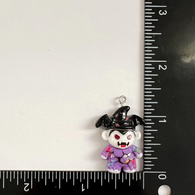 May include: A small vampire-themed charm featuring a white face with red eyes and fangs, wearing a black hat with bat wings. The charm has a purple body with a cracked pattern and colorful accents. A ruler shows it is approximately 1.25 inches tall.
