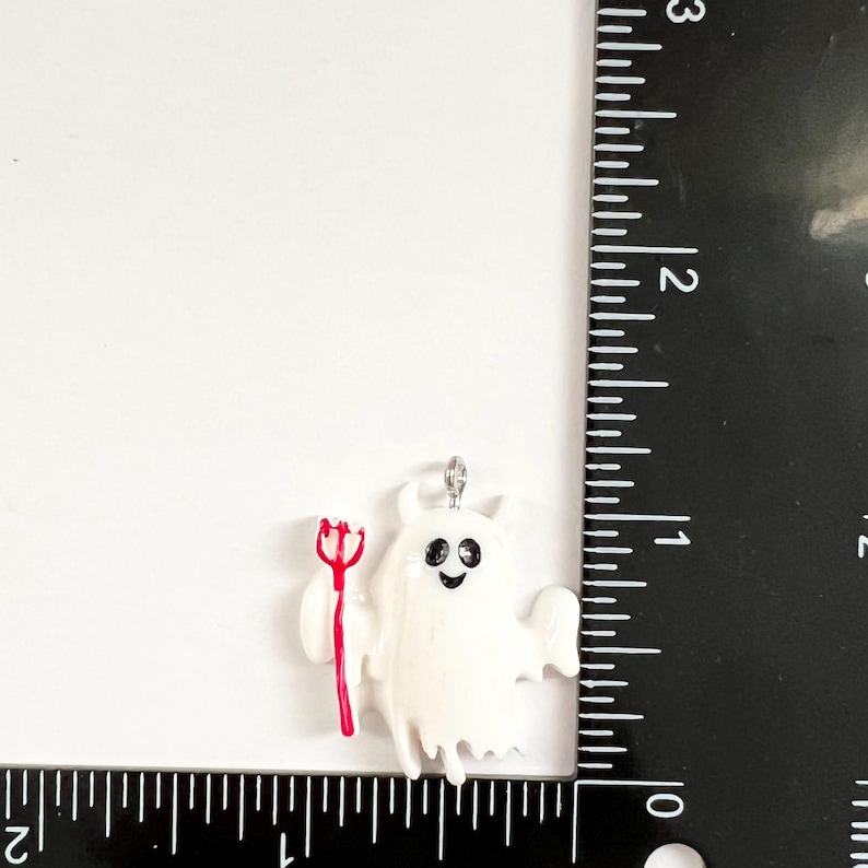 May include: A white ghost charm with black eyes and a smiling mouth, holding a red trident. The charm is positioned next to a black ruler, showing a height of approximately 1.25 inches and a width of approximately 1.25 inches.