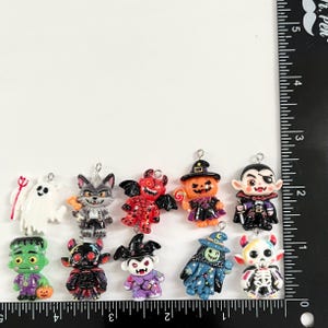 May include: A collection of ten Halloween-themed charms featuring a ghost, werewolf, devil, pumpkin witch, vampire, Frankenstein monster, horned creature, bat-winged figure, blue-robed witch, and skeleton, measured against a ruler showing 2.5 inches.