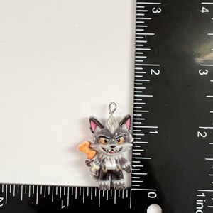 May include: A gray wolf charm with a white chest, pink inner ears, and orange eyes, holding an orange bone. The charm is positioned next to a black ruler, showing a height of approximately 1.25 inches and a width of approximately 1 inch.