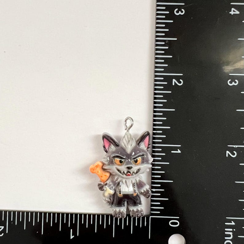 May include: A gray wolf charm with a white chest, pink inner ears, and orange eyes, holding an orange bone. The charm is positioned next to a black ruler, showing a height of approximately 1.25 inches and a width of approximately 1 inch.