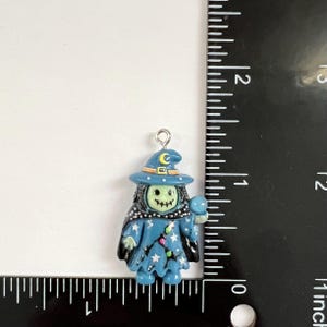 May include: A blue witch charm with a pale green face, wearing a hat with a yellow crescent moon and holding a blue orb. The charm features white star accents and a black cape. A ruler shows the charm is approximately 1.25 inches in height.