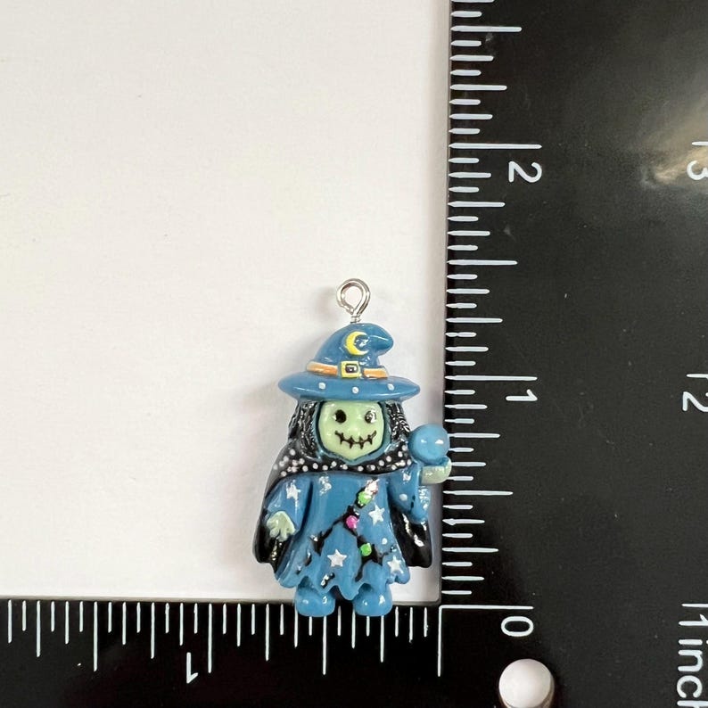 May include: A blue witch charm with a pale green face, wearing a hat with a yellow crescent moon and holding a blue orb. The charm features white star accents and a black cape. A ruler shows the charm is approximately 1.25 inches in height.