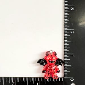 May include: A red and black devil charm with a small metal loop at the top. The figure features black wings, horns, and a textured body pattern. A black ruler is positioned along the side and bottom, showing the charm is approximately 1 inch tall.