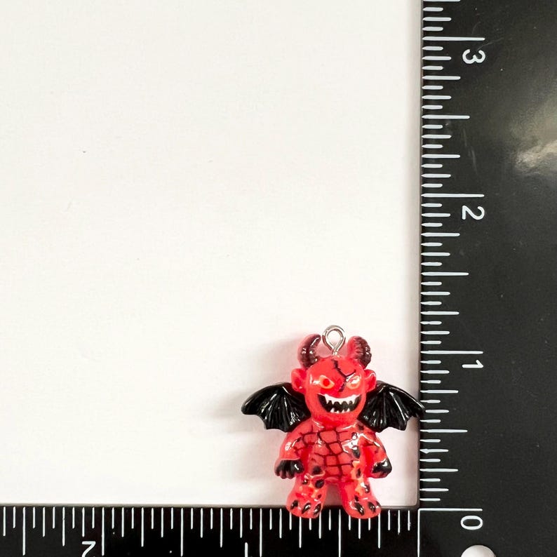 May include: A red and black devil charm with a small metal loop at the top. The figure features black wings, horns, and a textured body pattern. A black ruler is positioned along the side and bottom, showing the charm is approximately 1 inch tall.