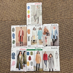 May include: A collection of eight Simplicity sewing patterns for coats and jackets, featuring various styles including hooded, belted, and open-front designs, displayed in white paper envelopes with printed illustrations of finished garments.