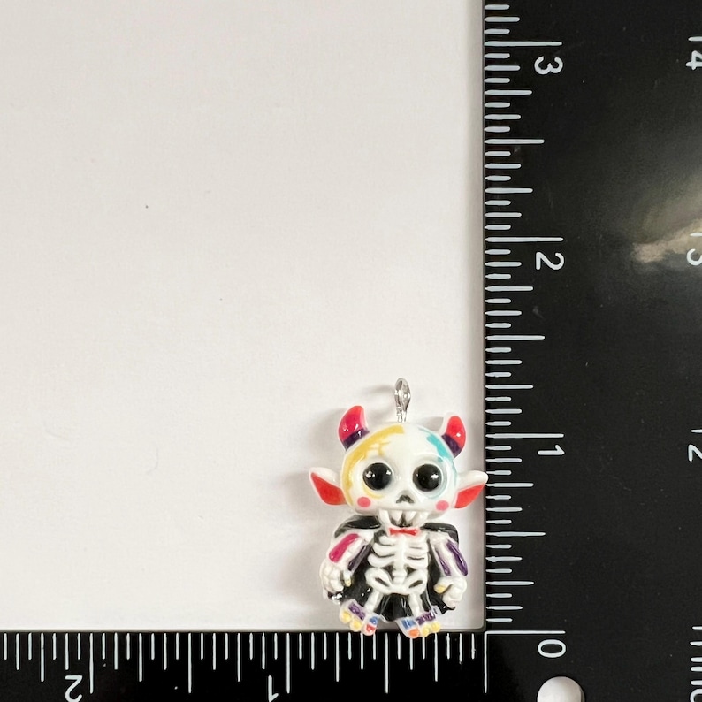 May include: A small, colorful skeleton charm with a white skull, black eyes, and red horns, positioned next to a black ruler showing a height of approximately 1.25 inches. The charm features a black cape and multi-colored accents on the head.