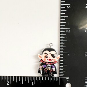 May include: A small vampire charm featuring a black cape, a white shirt with a red bowtie, and a black eye patch. The figure holds a silver sword and has a metal loop at the top. A black ruler shows the charm is approximately 1.25 inches tall.