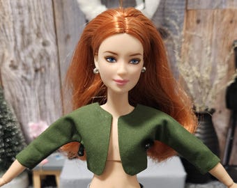 May include: A doll with red hair and blue eyes wearing a dark green bolero jacket. The doll has silver earrings and a neutral expression. The background includes a small Christmas tree and a black decorative mannequin.