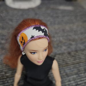 May include: A doll with red hair wearing a white fabric headband featuring a Halloween pattern of orange jack-o'-lanterns and black bats. The headband is finished with a purple stitched edge and is worn on the doll's head above a black top.