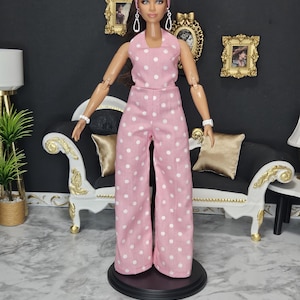 May include: A doll wearing a pink polka dot jumpsuit with a matching headband. The doll has long brown hair and is wearing silver earrings and a white bracelet. The doll is standing on a black base.