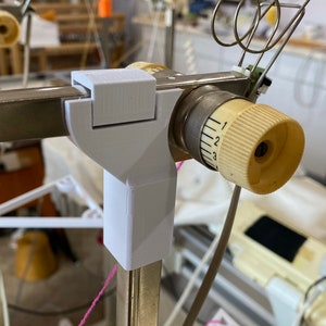 Op de afbeelding: Een witte, 3D-geprinte kunststof beugel bevestigd aan een metalen breimachine met een witte garenrol. De rol heeft een wijzerplaat met de nummers 1, 2 en 3.