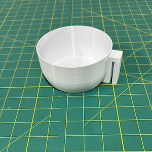 May include: A white 3D printed cup with a handle. The cup is on a green cutting mat.