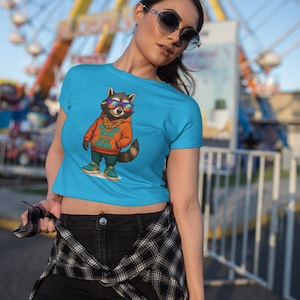 May include: A woman wearing a blue crop top with a cartoon raccoon wearing sunglasses and a gold chain. The raccoon is wearing an orange shirt with the text "ON THE GRID IN MY GRIND". The woman is wearing black shorts and a plaid shirt tied around her waist.