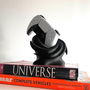 May include: A silver and black games controller sits on a black, swirling stand. The controller is placed on top of two books, one titled "UNIVERSE" and the other "STAR WARS COMPLETE VEHICLES". A black, abstract sculpture is to the right.