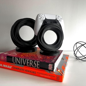 May include: A white and black PlayStation 5 controller sits in a black circular stand. Another black circular stand is next to it. The stands are on top of two books titled "UNIVERSE" and "STAR WARS COMPLETE VEHICLES". A black wire sculpture is to the right.