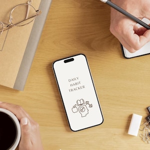 Op de afbeelding: Een witte smartphone met een zwart scherm waarop een app voor het bijhouden van dagelijkse gewoontes wordt weergegeven. De app toont een vinkje-icoon, een boek en een halter. De tekst op het scherm luidt "Daily Habit Tracker".