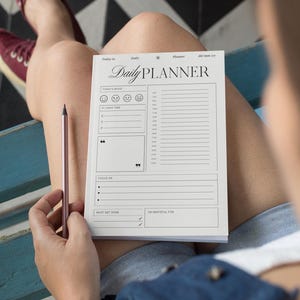 Op de afbeelding: Een witte dagelijkse planner met een zwart-wit ontwerp. De planner heeft secties voor "Humeur van vandaag", "Te doen", "Focus op", "Moet gedaan worden" en "Ik ben dankbaar voor".
