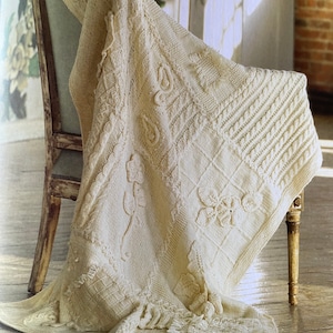 May include: Cream-colored knitted blanket draped over a wooden chair. The blanket features various textured patterns, including floral and geometric designs. The blanket's edge has a fringe detail, adding a decorative touch to the cozy home decor item.