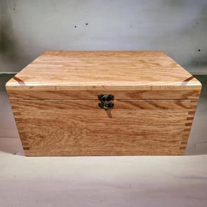 May include: A wooden box with a hinged lid and a metal clasp. The box is made of light-coloured wood with visible grain and dovetail joints. The box is rectangular and appears to be empty.