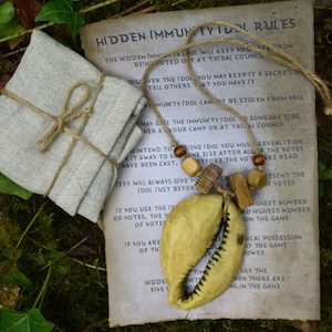 May include: A wooden and shell necklace with a set of rules for a hidden immunity idol game. The rules are written on a piece of paper with a brown background. The necklace is made of a variety of materials, including wood, shell, and beads. The necklace is tied with a brown string.