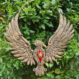 May include: A golden-colored, stylized bird sculpture with outstretched wings and red accents. The bird is set against a backdrop of green foliage. The sculpture features detailed feathering and a central red emblem.
