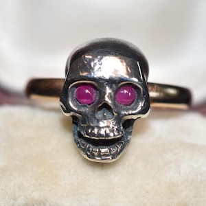 Puede incluir: Un anillo de plata con forma de calavera con dos ojos de piedras preciosas rosas.