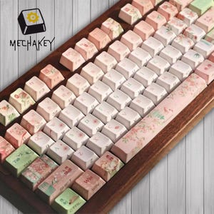 May include: A mechanical keyboard with a dark wood frame and custom keycaps featuring floral patterns in shades of pink, white, and green. The keys display letters, numbers, and symbols, with some keys showing Asian characters. The logo MECHAKEY is visible.