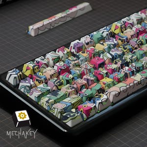 May include: A mechanical keyboard with vibrant, cartoon-style keycaps. The keycaps display abstract designs in pink, green, yellow, and blue. A matching spacebar keycap is also present. The brand logo "MECHAKEY" is visible in the bottom left corner.