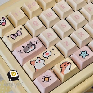 May include: A keyboard with beige and pink keycaps. Some keycaps feature pixelated cat and bow designs. The keys have letters and numbers in a pink font. The brand name "MECHAKEY" is visible on a black and yellow logo.
