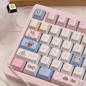 May include: A pink mechanical keyboard with custom keycaps featuring cartoon animal designs. The keyboard has white, pink, and blue keys with various symbols and letters. The brand name "MECHAKEY" is visible.