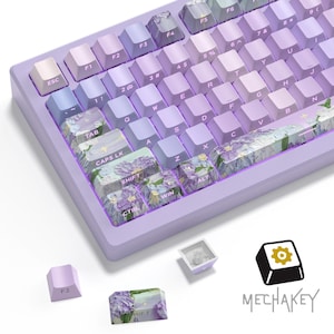 Könnte beinhalten: Eine lavendelfarbene mechanische Tastatur mit Keycaps in Lila- und Blautönen. Einige Tasten haben Blumendesigns. Die Tastatur hat einen hellvioletten Rahmen und der Markenname "MECHAKEY" ist sichtbar.