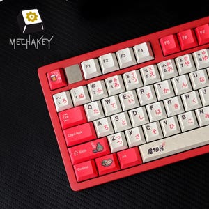 May include: A red and white mechanical keyboard with Japanese characters and custom keycaps. The keyboard has a red frame, white keys with Japanese characters, and custom keycaps with illustrations. The brand name "MECHAKEY" is visible in the upper left.